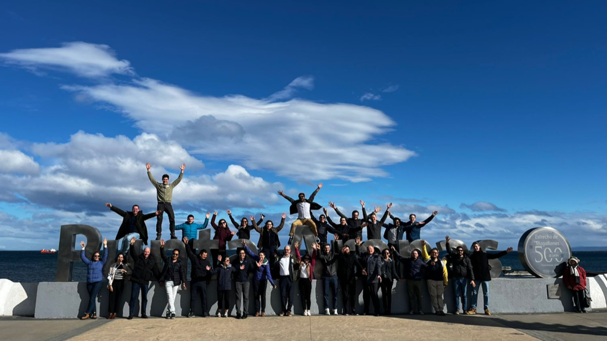 Group of people posing on large metal letters "Punta Arenas" with ocean backdrop under a bright blue sky.